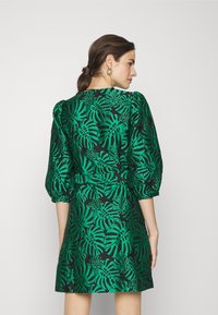 Robe verte avec imprimé de feuilles noires, présentant des manches bouffantes et une taille cintrée, fabriquée en tissu lisse. Vue de dos, design à mi-genoux.