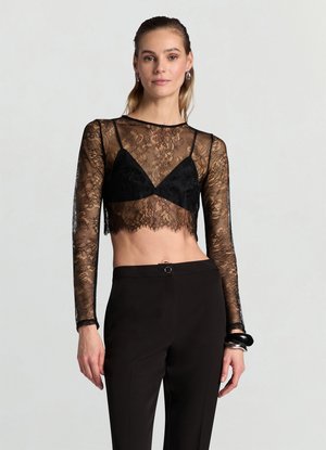Top corto in pizzo nero con maniche lunghe, indossato sopra un reggiseno nero, caratterizzato da un orlo smerlato e una texture trasparente. Abbinato a pantaloni neri.