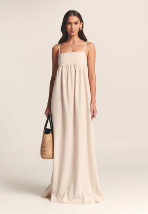 Maxikleid aus leichtem beigen Leinen mit dünnen Trägern, gerafftem Bund und fließender Silhouette. Begleitet von einer geflochtenen Strohtasche.