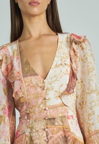 Kobieta nosząca różową, beżową i kremową sukienkę w kwiatowy wzór z falbany na ramionach i guzikami pokrytymi materiałem z przodu.