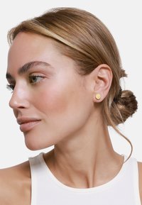 Goldene Steckohrringe mit strukturiertem Finish, runder Form und minimalistischem Design, getragen von einem Modell mit glatter Haut und hellen Haaren.
