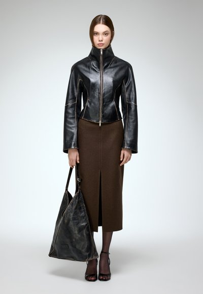 Chaqueta de cuero negra con cuello alto y cremallera plateada, combinada con una falda marrón hasta la rodilla con una abertura frontal, llevando un gran bolso de cuero negro.