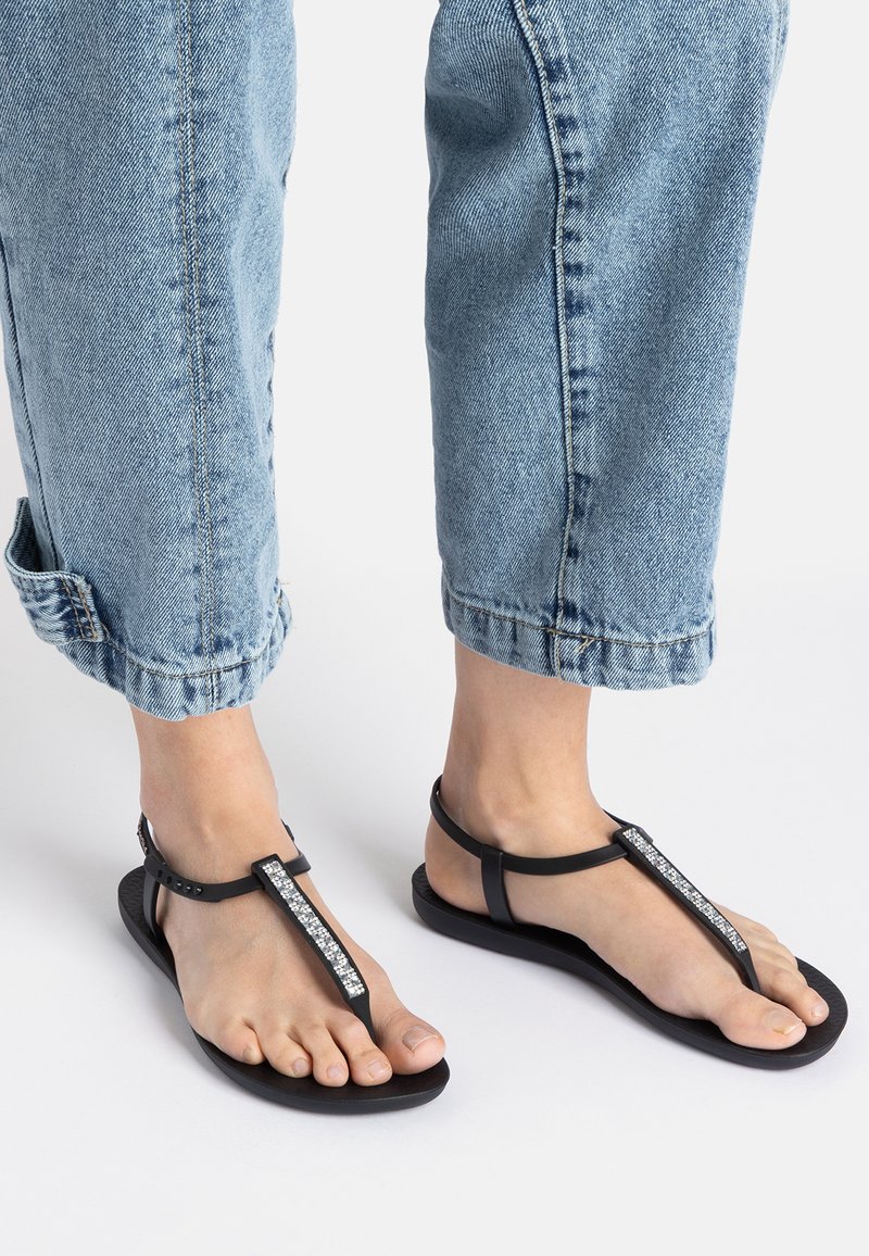 Schwarze Sandalen mit einem Riemen, der zwischen den Zehen verläuft, und einem Strassakzent an der Oberseite. Die Sohle ist flach und aus Gummi. Getragen mit umgeschlagenen Jeans.