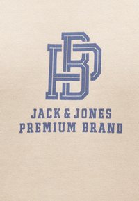 Beige stof met een getextureerd oppervlak; bevat een blauw gedrukt logo "HB" en "JACK & JONES PREMIUM BRAND" in vetgedrukte letters.