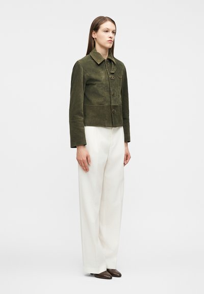Γυναίκα που στέκεται φορώντας ένα κλειστό σουέτ τζάκετ σε ελιά πράσινο χρώμ α, άσπρο παντελόνι με φαρδιά πόδια και καφέ μυτερά παπούτσια, μπροστά σε έναν απλό λευκό φόντο.
