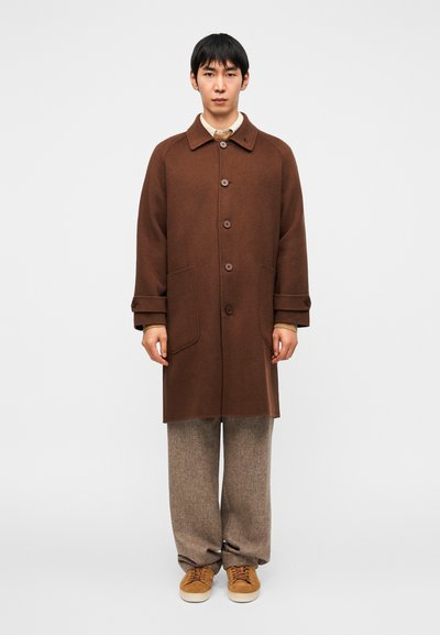 Abrigo marrón con cuello, botones delanteros y bolsillos laterales, combinado con pantalones de cuadros marrón claro y zapatillas marrón claro. Diseño sencillo y estructurado.