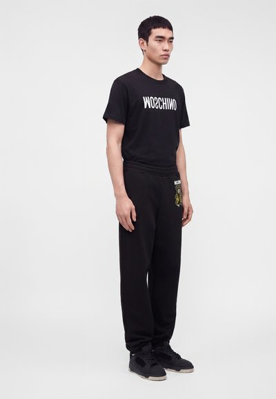 Νέος άνδρας με μαύρο t-shirt Moschino και μαύρη φόρμα με κέντημα αρκούδας, στέκεται μπροστά σε απλό λευκό φόντο.