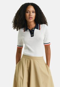 Camisa polo blanca con relieve, cuello azul marino y acentos rojos, con botones dorados. Combinada con una falda acampanada beige.