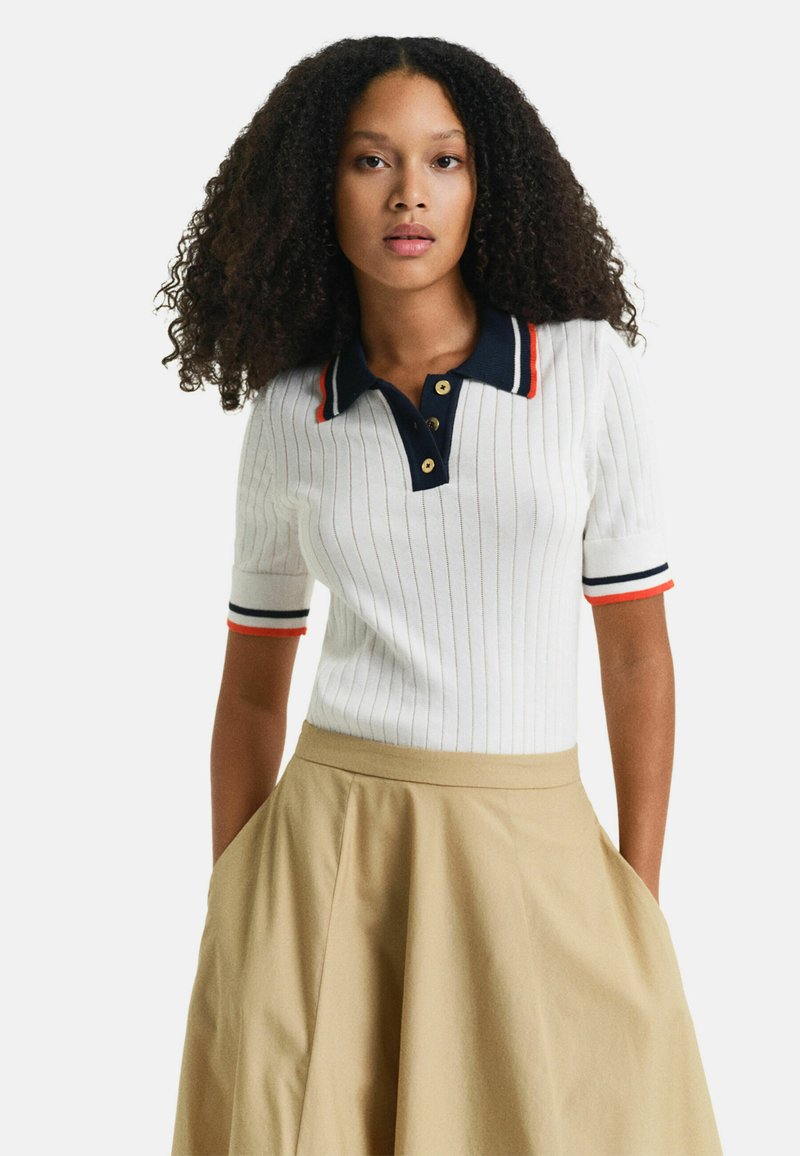 Camisa polo blanca con relieve, cuello azul marino y acentos rojos, con botones dorados. Combinada con una falda acampanada beige.