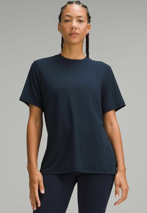 Femme aux cheveux tressés portant un T-shirt à manches courtes bleu marine uni et un pantalon assorti, debout devant un fond neutre.