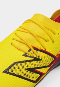 Κοντινό πλάνο ενός φωτεινού κίτρινου αθλητικού παπουτσιού με κίτρινα κορδόνια και μεγάλο μαύρο και κόκκινο στυλιζαρισμένο λογότυπο "N" στο πλάι.