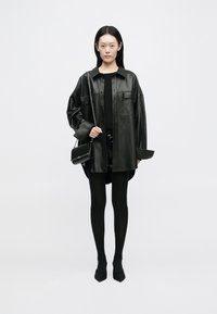 Giacca camicia in pelle nera oversize con due tasche frontali, indossata sopra un top nero e una mini gonna. Leggings neri e scarpe a punta completano il look.