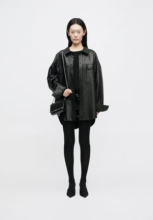 Giacca camicia in pelle nera oversize con due tasche frontali, indossata sopra un top nero e una mini gonna. Leggings neri e scarpe a punta completano il look.