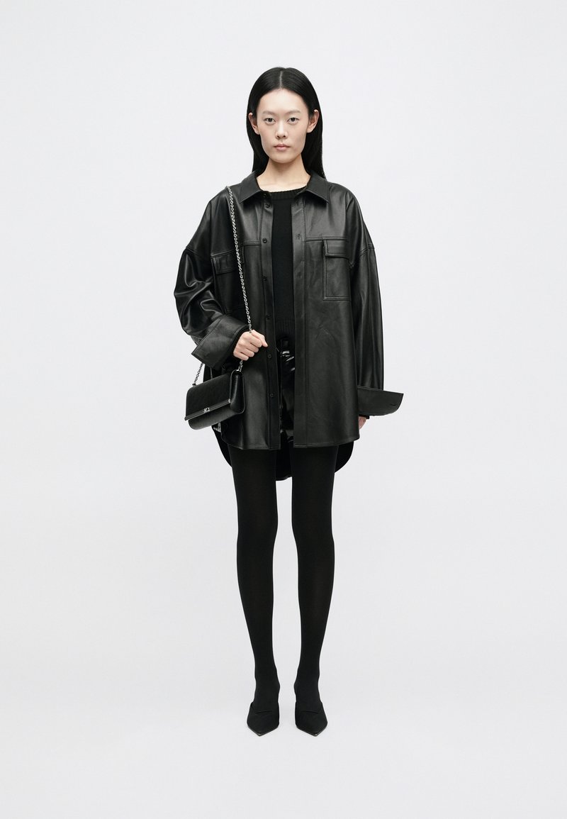 Giacca camicia in pelle nera oversize con due tasche frontali, indossata sopra un top nero e una mini gonna. Leggings neri e scarpe a punta completano il look.