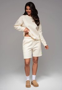Ensemble en sweat-shirt et shorts couleur crème, fabriqué dans un tissu doux, comportant une taille ajustable et un petit accent logo vert.