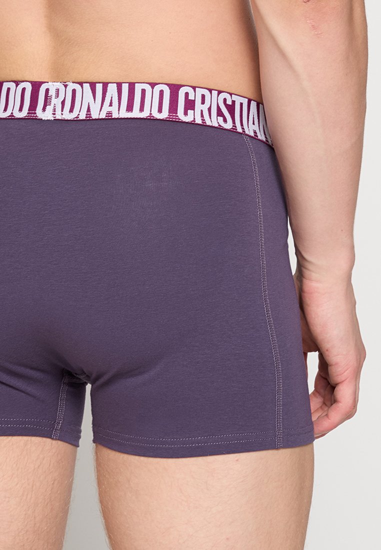 Boxer corto morado con una banda blanca que presenta el texto "Cristiano Ronaldo". La tela parece suave con una textura lisa y un ajuste ceñido.