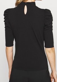 Top aderente nero realizzato in tessuto morbido, con maniche a sbuffo, collo alto con una piccola apertura a forma di chiave sul retro e maniche corte.