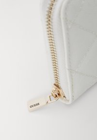 Portefeuille matelassé blanc avec une fermeture éclair dorée et une étiquette gravée "GUESS". Présente une surface texturée et une forme rectangulaire élégante.