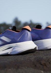 Lilla sportsko med orange accent, der har en tekstureret overdel, tre hvide striber og en hvid mellemsål med branddetaljer.