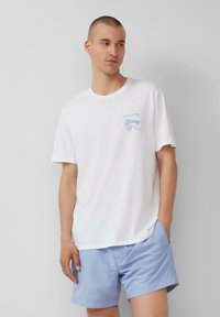 Weißes Baumwoll-T-Shirt mit Rundhalsausschnitt und einem blauen Druckmotiv auf der Brust. Getragen mit hellblauen Shorts. Glatte Textur.