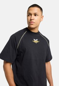 Sort t-shirt med rund halsudskæring, korte ærmer og kontrasterende gule syninger langs skuldrene. Inkluderer et broderet logo-design.