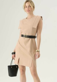Beige šaty s volánky na ramenou, bočními rozparky a černým páskem se zlatou sponou. Modelka drží malou černou kabelku.