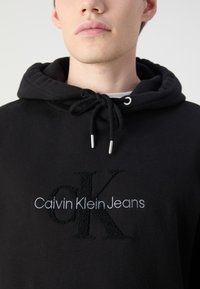 Zwarte katoenen hoodie met een trekkoord in de capuchon, een geborduurd logo aan de voorkant in textuur zwart en grijs, en een standaard pasvorm.