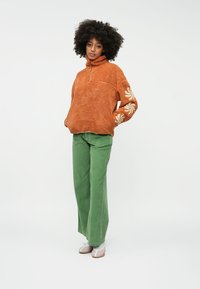 Pulôver de fleece castanho com fecho de meia-zíper e bordado floral na manga, combinado com calças largas de veludo cotelê verdes e calçado claro.