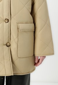 Krémová prošívaná péřová bunda s knoflíkovým zapínáním, boční kapsou a širokými rukávy. Hladká textura a detail v diamantech.