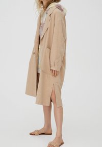 Persona con abrigo beige hasta las rodillas y una abertura lateral, sobre una sudadera con capucha de tonalidad pastel y sandalias trenzadas en color marrón claro, frente a un fondo liso.