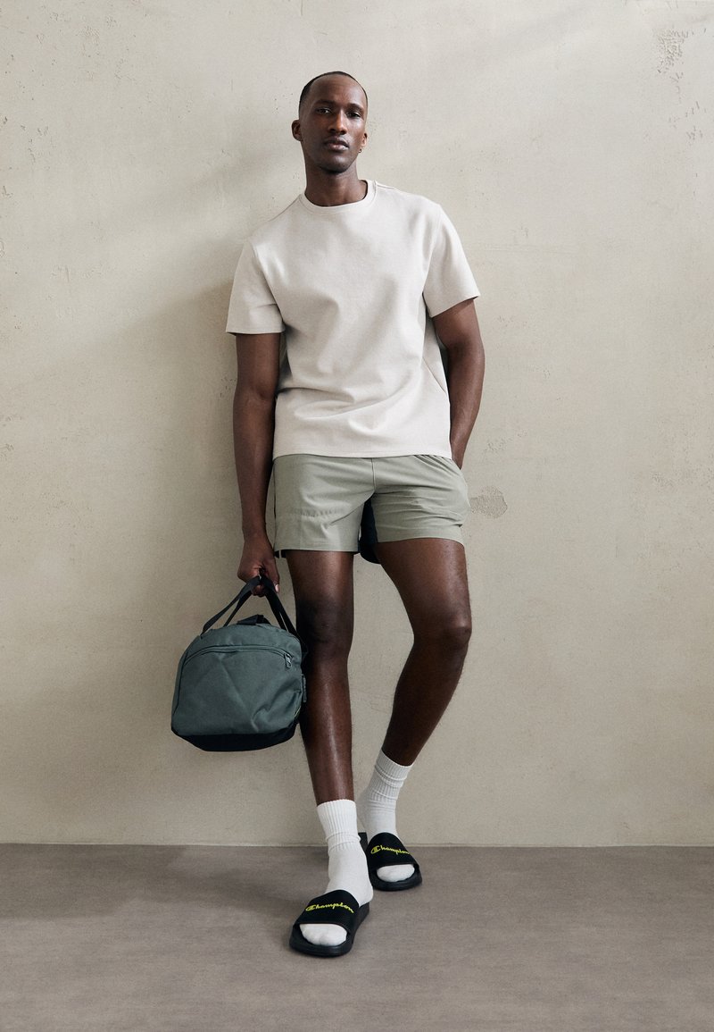 Ein schlichter beiger T-Shirt, hellgrüne Shorts, weiße Socken, schwarze Sandalen mit gelbem Logo, hält eine dunkelgrüne Tasche vor einem neutralen Hintergrund.