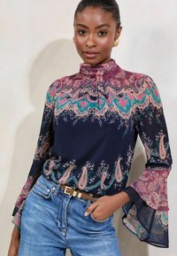 Mornarica bluza z visokim nabranim ovratnikom, paisley vzorcem v teal, roza in krem barvah, ter z razširjenimi prosojnimi rokavi, v kombinaciji z modrimi kavbojkami in rjavim pasom.