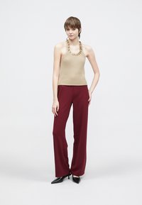 Beige geribbelde haltertop met gerafelde halslijn, gecombineerd met losvallende bordeauxrode broek en zwarte schoenen met puntige neus.