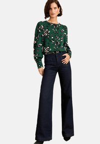 Groene bloemenblouse met pofmouwen, gecombineerd met donkerblauwe wijdbenige jeans. Blouse heeft knopen en een ontspannen pasvorm.