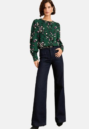 Blusa floreale verde con maniche a sbuffo, abbinata a jeans a gamba larga di colore blu scuro. La blusa presenta bottoni e una vestibilità comoda.