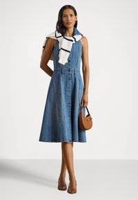 Džinsa kleita ar pogām priekšpusē, bez piedurknēm un ar izplešamu svārku. Balts džemperis, sasiets ap kaklu. Brūna rokassoma.