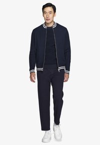 Marineblå bomberjakke med teksturerede strikkede ærmer og hvide stribede ribkantbånd. Båret over en mørk sweater, parret med mørke bukser og hvide sneakers.