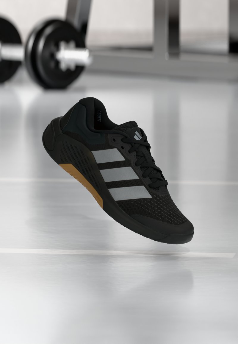 Μαύρο αθλητικό παπούτσι με πλέγμα στην επάνω επιφάνεια, τρεις ασημένιες ρίγες και λαστιχένια σόλα με υφή για καλύτερο κράτημα. Το παπούτσι αιωρείται στο χώρο με φόντο ένα γυμναστήριο.