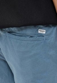 Pantaloni blu chiaro con una texture liscia, dotati di una tasca posteriore e di un'etichetta in tessuto bianco con la scritta "JACK & JONES EST. 1990." Top nero a costine visibile sopra.