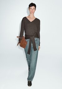 Brauner Wickeloberteil mit einem Bindegürtel in der Taille, kombiniert mit hellblauen, hoch taillierten Jeans. Eine braune Lederhandtasche und schwarze Schuhe runden den Look ab.