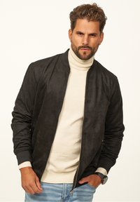 Schwarze Wildleder-Bomberjacke mit gerippten Bündchen und Kragen, mit Reißverschluss, getragen über einem cremefarbenen Rollkragenpullover. Lässiger, figurbetonter Stil.