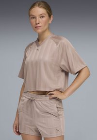 Γυναίκα που φοράει ροζ αθλητικό crop top Puma και ταιριαστά σορτς, στέκεται με το ένα χέρι στη μέση μπροστά από απλό γκρι φόντο.