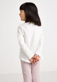 Ragazzina con capelli scuri e lisci, sorridente, rivolta a sinistra, indossa una maglietta bianca a maniche lunghe e pantaloni rosa chiaro, con le mani unite dietro la schiena.