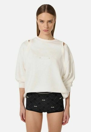 Donna con capelli lisci indossa una felpa bianca oversize con spacchi sulle spalle e pantaloncini neri con fantasia, in piedi contro uno sfondo semplice.