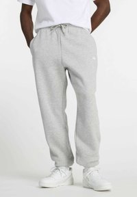 Muž v svetlosivých teplákoch s šnúrkou a bielych teniskách, stojaci s rukami v vreckách na jednom pozadí.