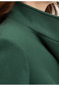 Grønt tekstureret stof med en struktureret krave og glat finish, der fremhæver syningsdetaljerne og de rene linjer i designet.