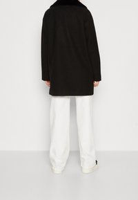 Svart kappa med krage av syntetisk päls, långa ärmar och lös passform. Kombinerad med vita byxor med vida ben och ljusa skor.