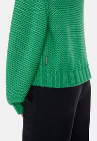 Grüner gestrickter Pullover mit strukturiertem Muster, weiten Bündchen und geripptem Saum. Enthält ein kleines Markenetikett an der Rückseite.