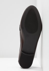 Semelle d'une chaussure en cuir marron foncé indiquant la taille 37, appuyée verticalement contre un mur blanc sur un sol blanc.