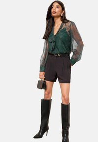 Mørk grøn gennemsigtig bluse med knyttet halsudskæring, parret med sorte high-waisted shorts og sort knæhøjhælede støvler. Lille sort håndtaske inkluderet.